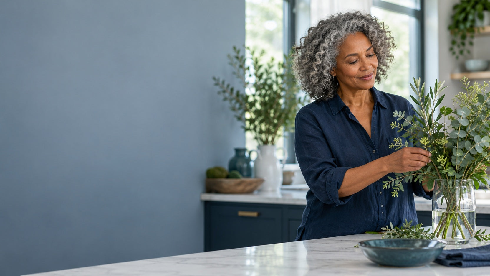 A woman arranging green foliage in a sunlit kitchen